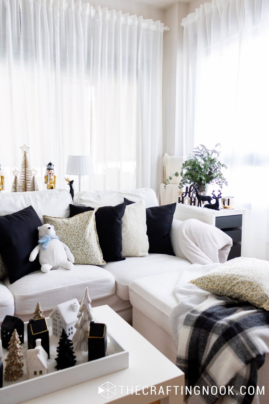 White Christmas polar bear with a blue scarf sitting among black and gold pillows on a white sectional sofa.
