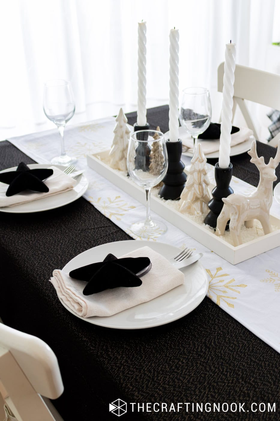 Close-up of white dinner plates with black star napkin decorations, gold snowflake runner, and Christmas centerpiece in a white tray.
