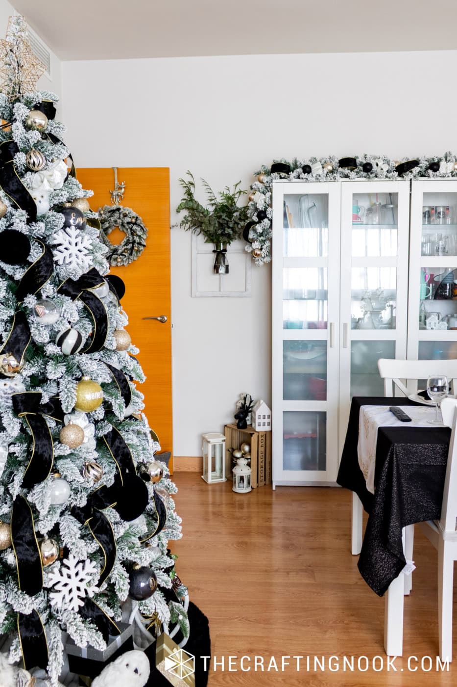 Flocked Christmas tree with black velvet ribbon beside white dining room cabinets decorated with matching snowy garland.
