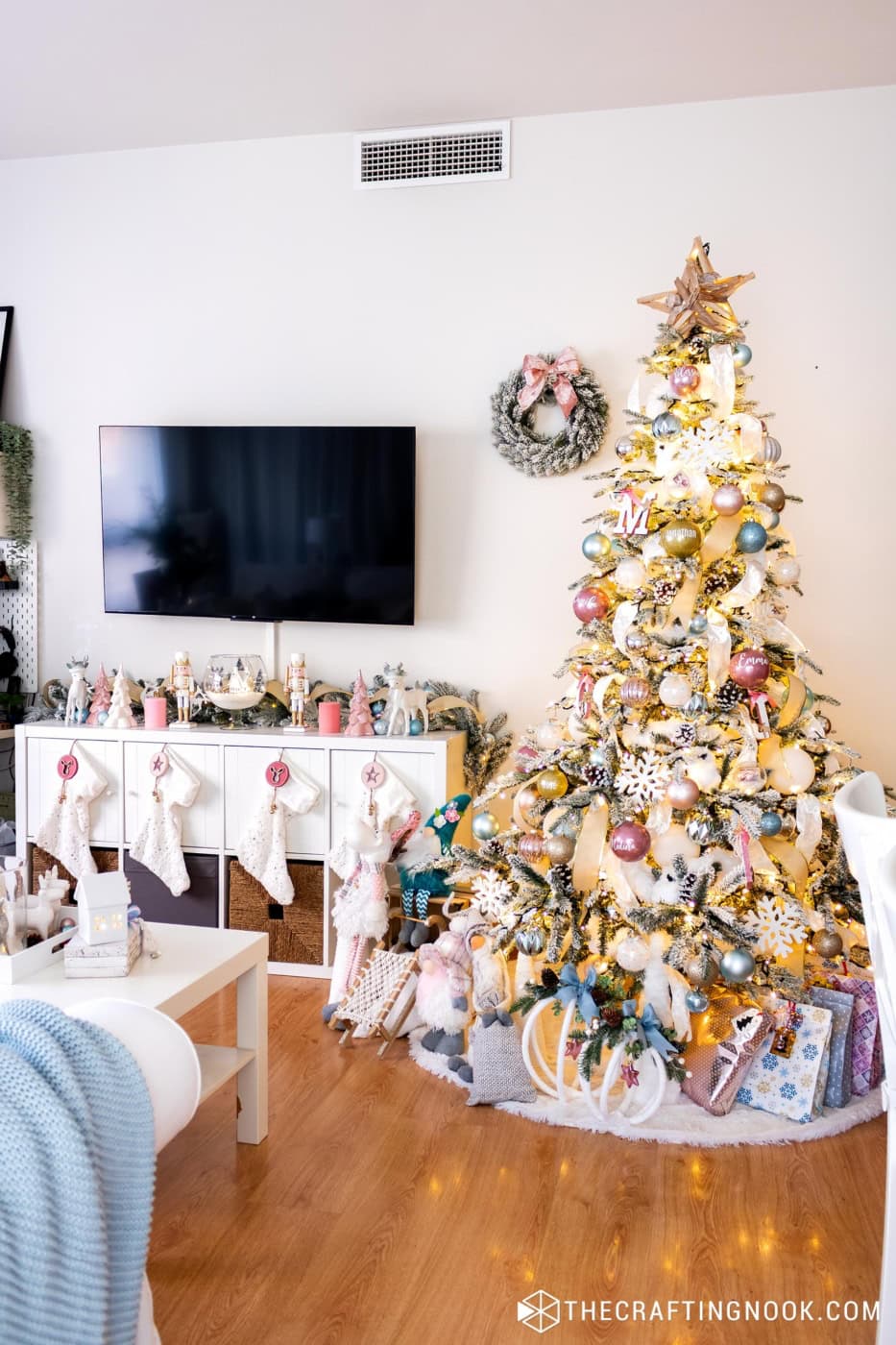 Flocked Christmas tree with pink, blue and gold ornaments next to a TV console with white stockings