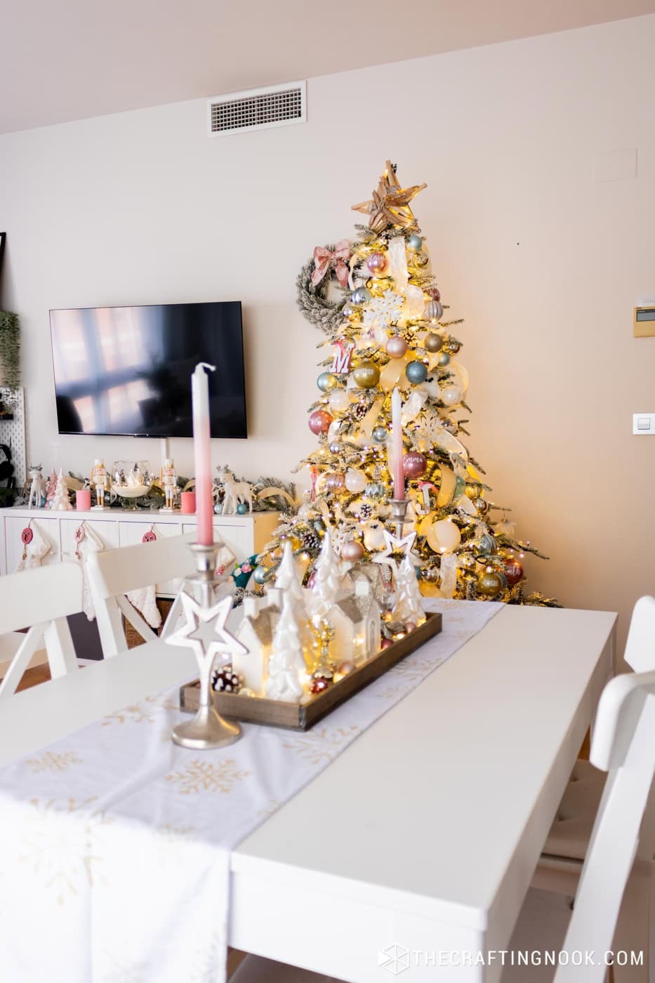Dining table with a snowflake runner and village centerpiece, with a lit pastel Christmas tree and TV console behind it