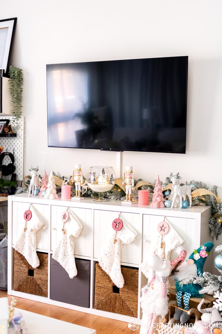 White shelf under a TV decorated with nutcrackers, reindeer, pink trees and four fluffy stockings
