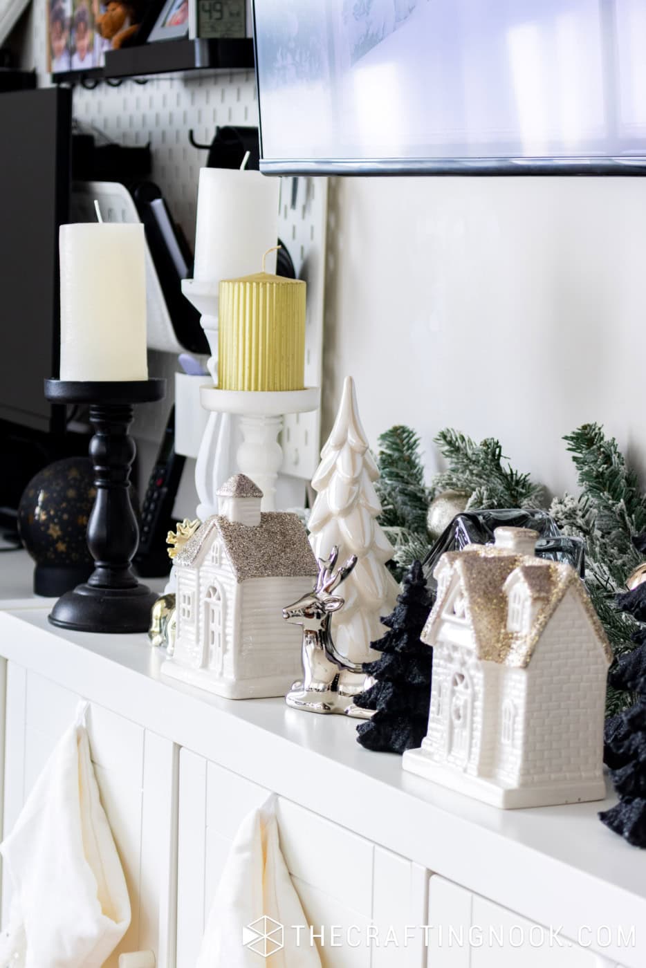 White Christmas TV console decorated with ceramic houses, black and white candleholders, gold candles, snowy garland, and metallic reindeer.
