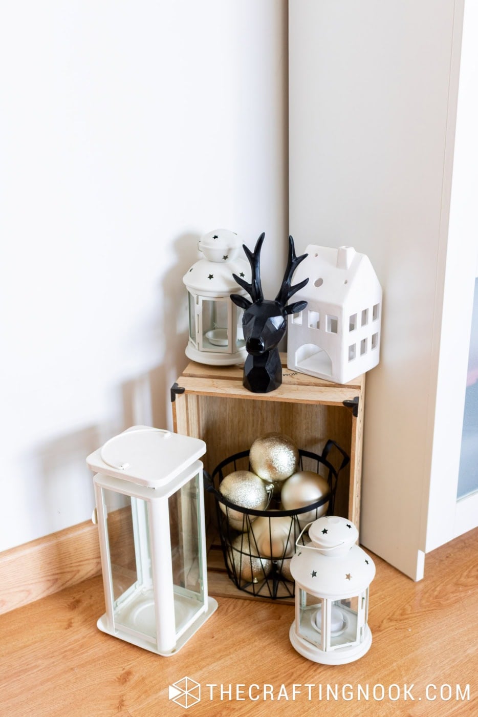 Christmas corner display with white lanterns, black reindeer figurine, ceramic house, and gold ornaments in a wire basket.
