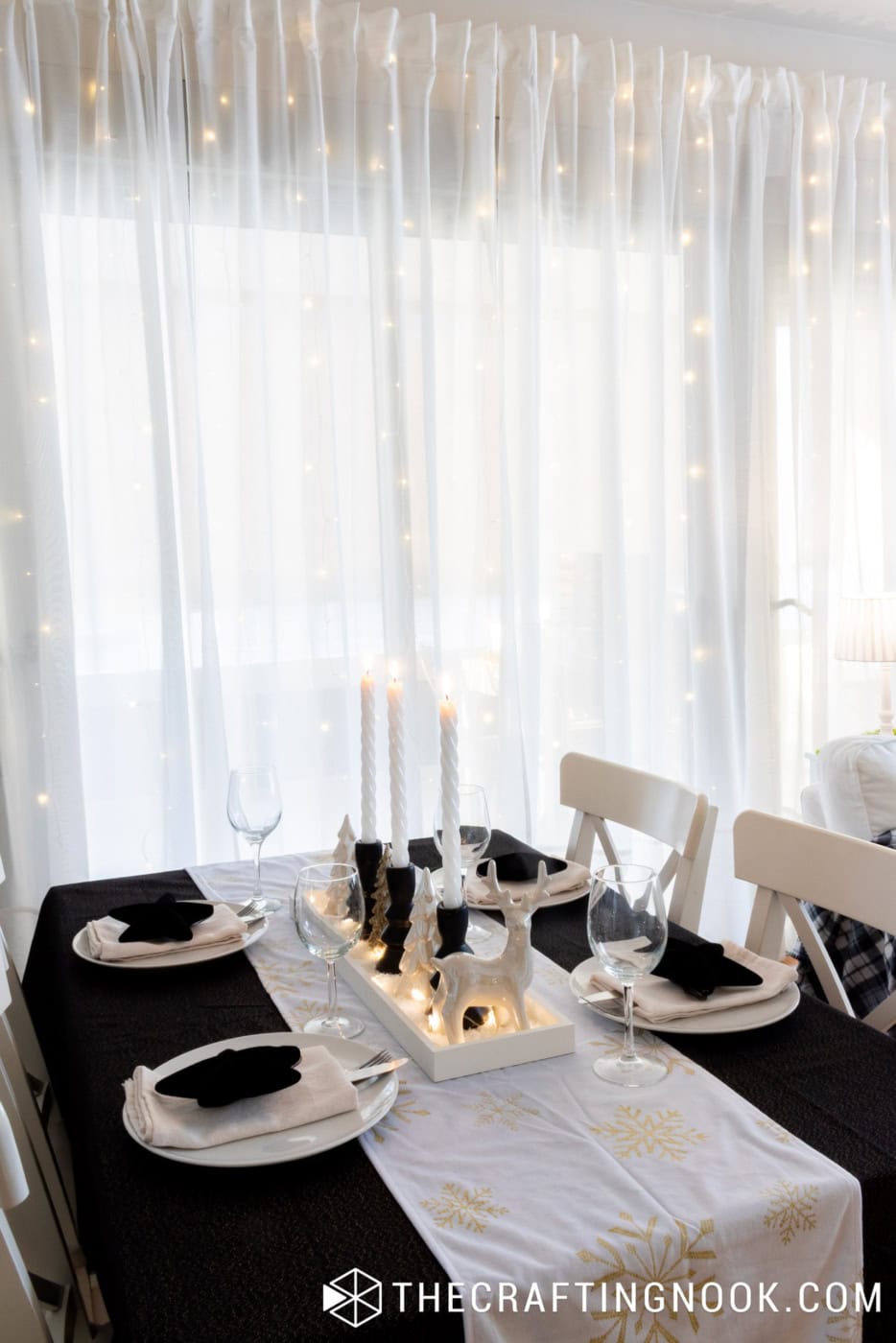 Black and white Christmas dining table with glowing candles, illuminated centerpiece, and warm fairy lights behind sheer curtains.
