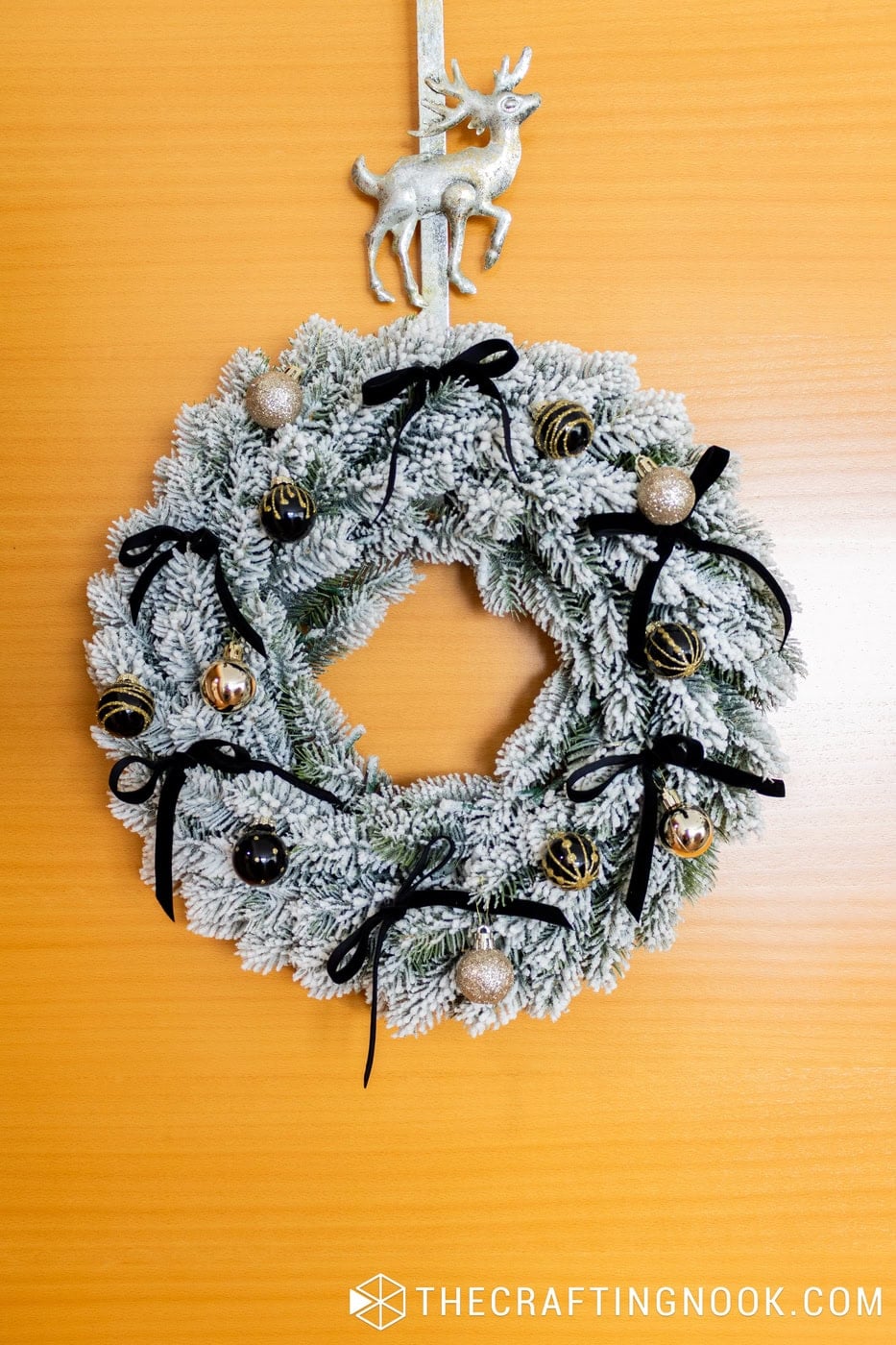 Snowy Christmas wreath with black ribbon bows and gold ornaments hanging on a wooden front door beneath a silver reindeer.
