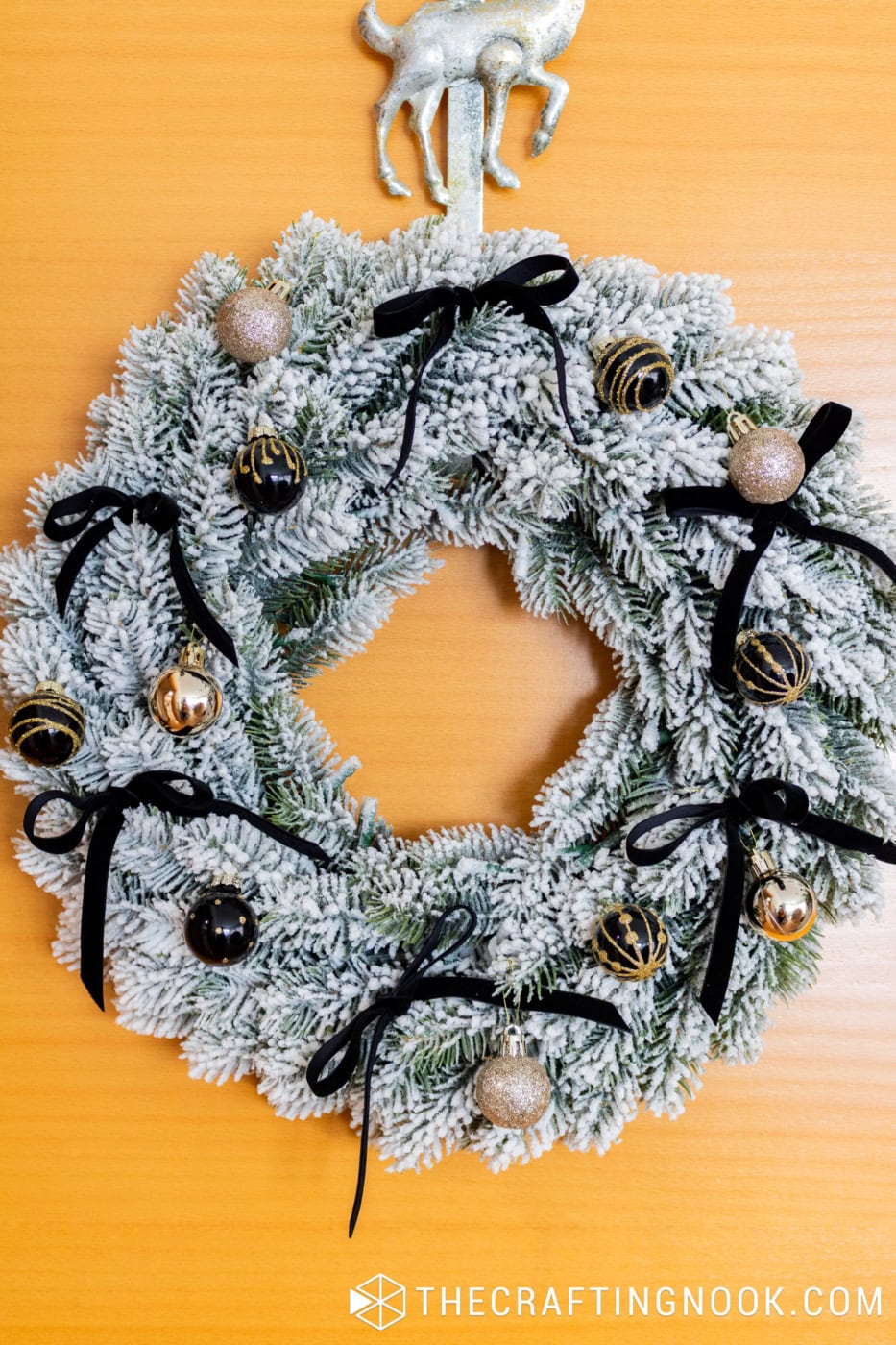 Flocked Christmas wreath decorated with black velvet bows, black and gold ornaments, and a silver reindeer door hanger.
