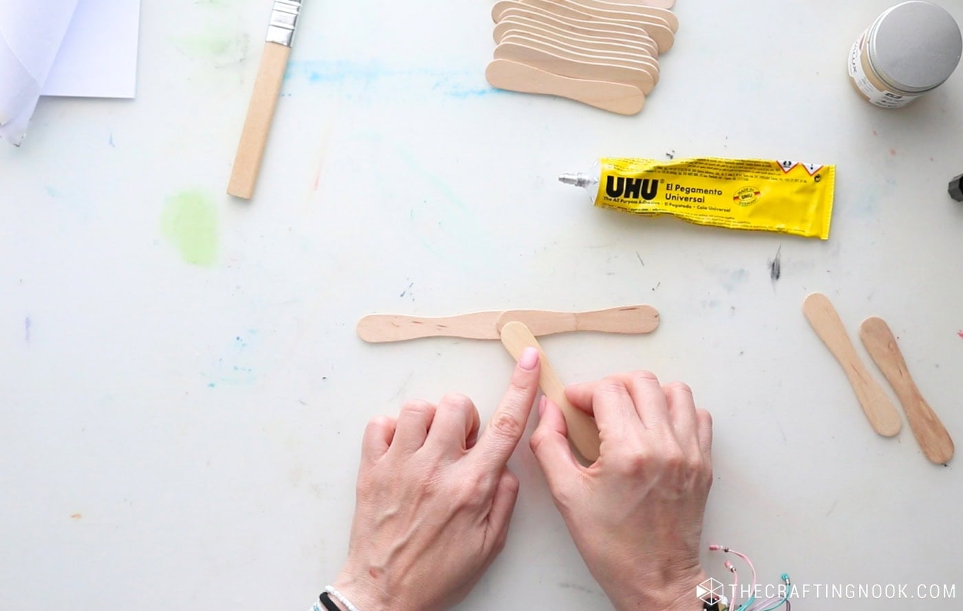 Gluing two wooden popsicle sticks together at the center to begin making a DIY snowflake.