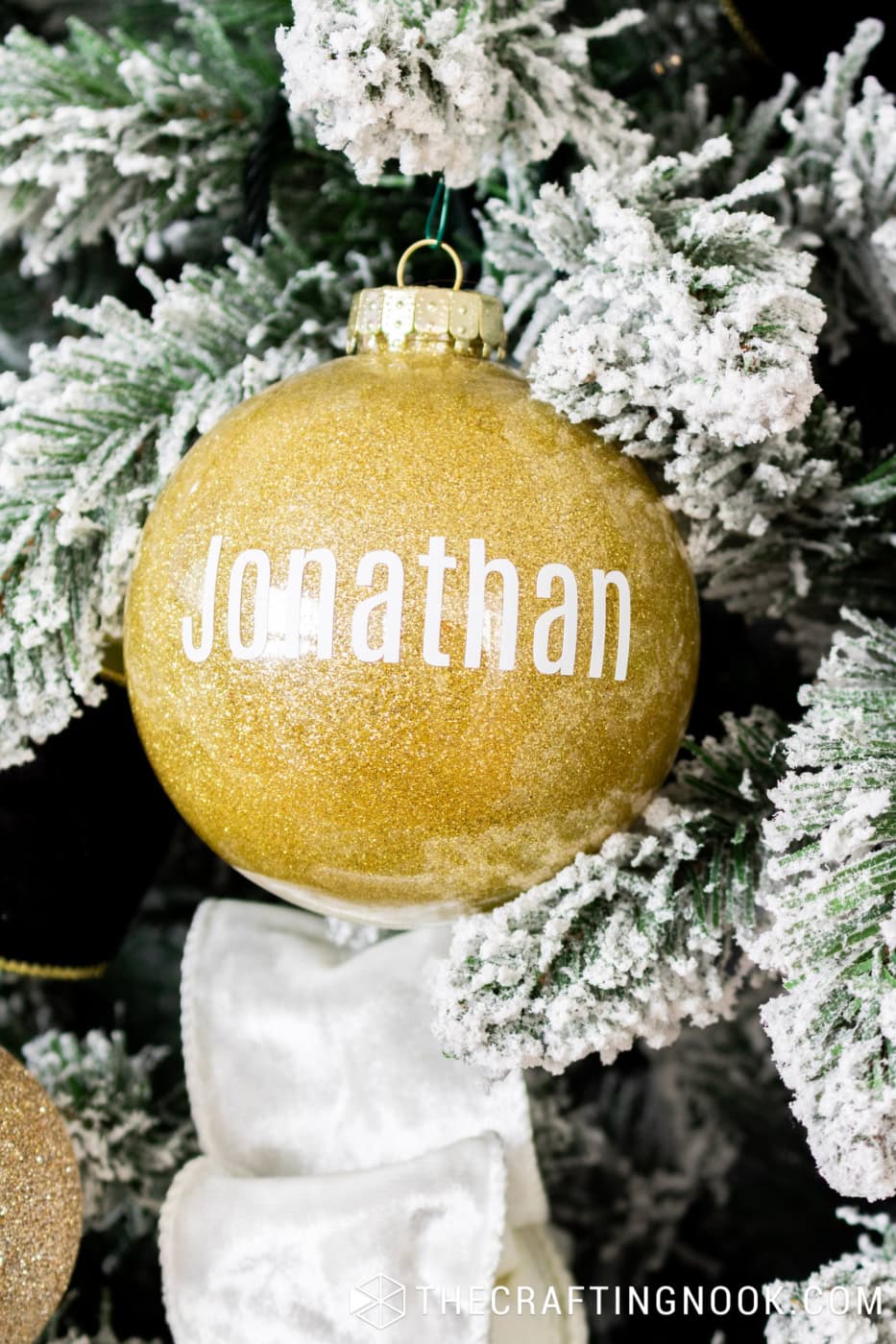 Gold glitter Christmas ornament with Jonathan's name in white lettering hanging from snow-covered Christmas tree branches.