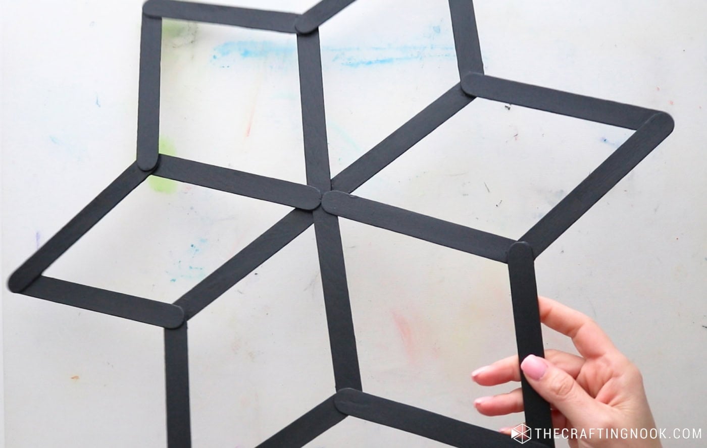 Holding a completed black popsicle stick snowflake with six open diamond-shaped points.
