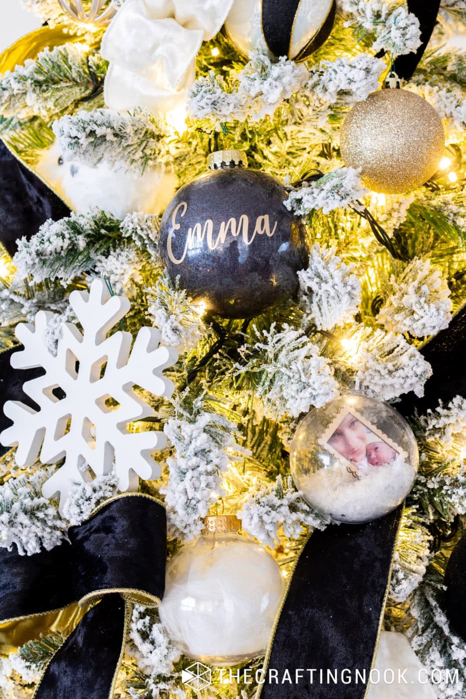 Personalized black glitter ornament, clear photo keepsake ornament and white snowflake surrounded by snowy branches and warm Christmas lights.