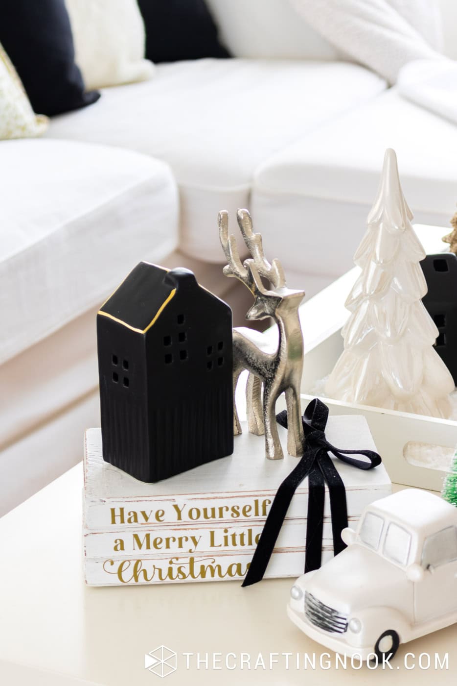 Handmade white Christmas stacked books with gold lettering and black ribbon, topped with a black ceramic house and silver reindeer beside a white pickup truck.
