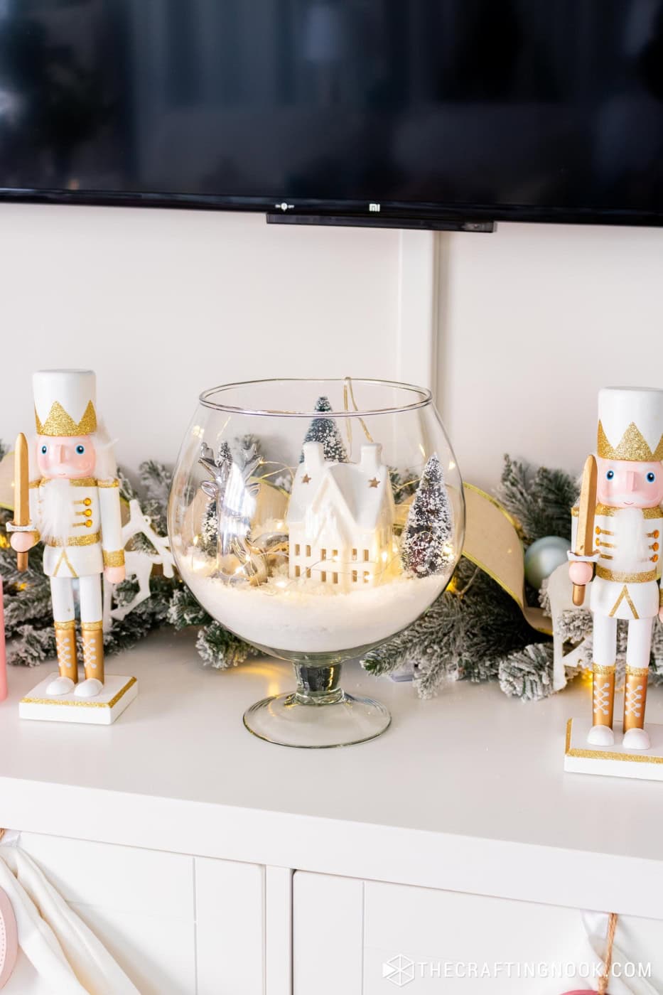 Large glass bowl with fake snow, a lit white house, snowy trees and a silver reindeer, between two white and gold nutcrackers