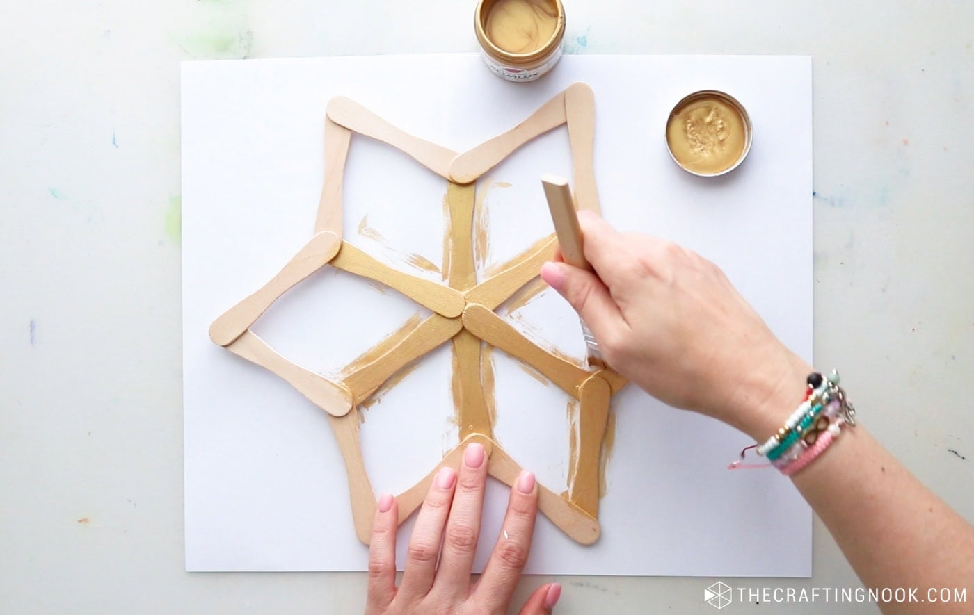 Painting a handmade popsicle stick snowflake with metallic gold acrylic paint and a brush.