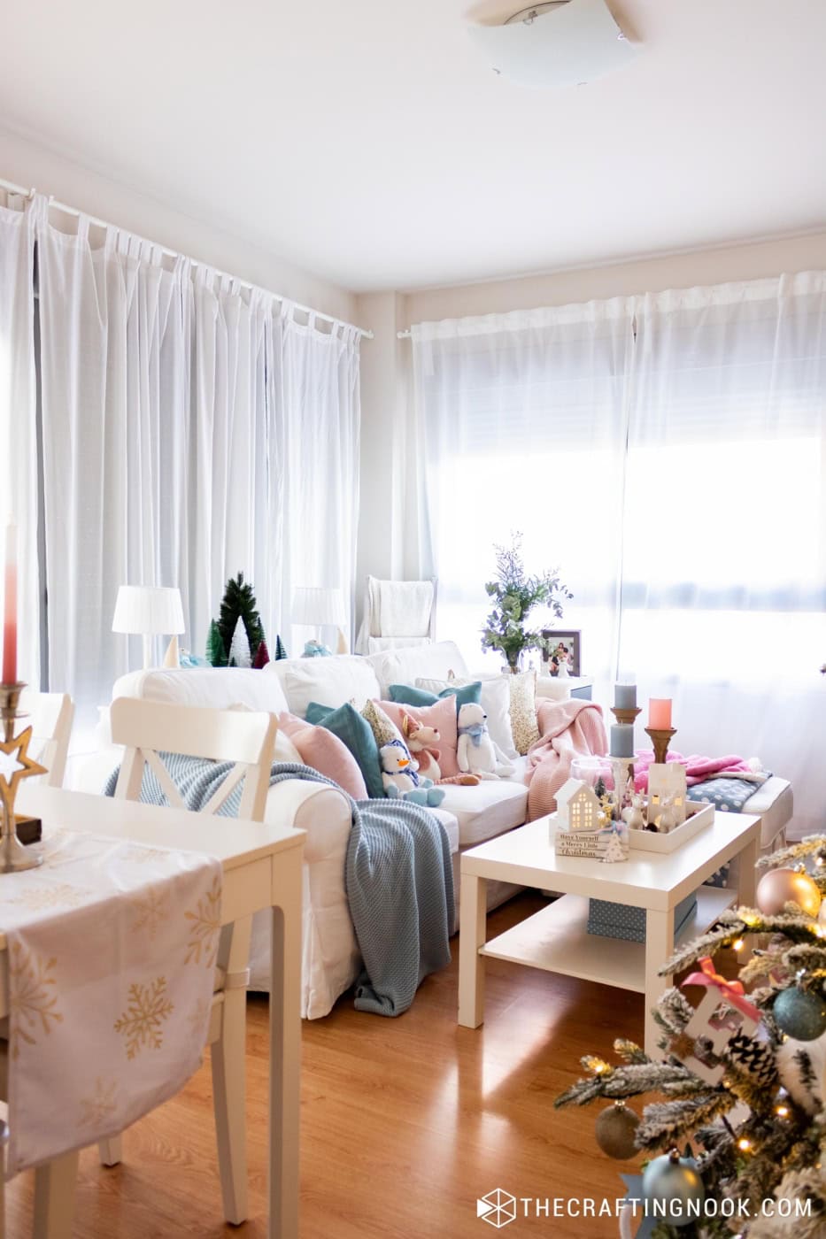 White sofa with pink and blue pillows and blankets in a bright living room decorated for Christmas, with a lit tree in the corner