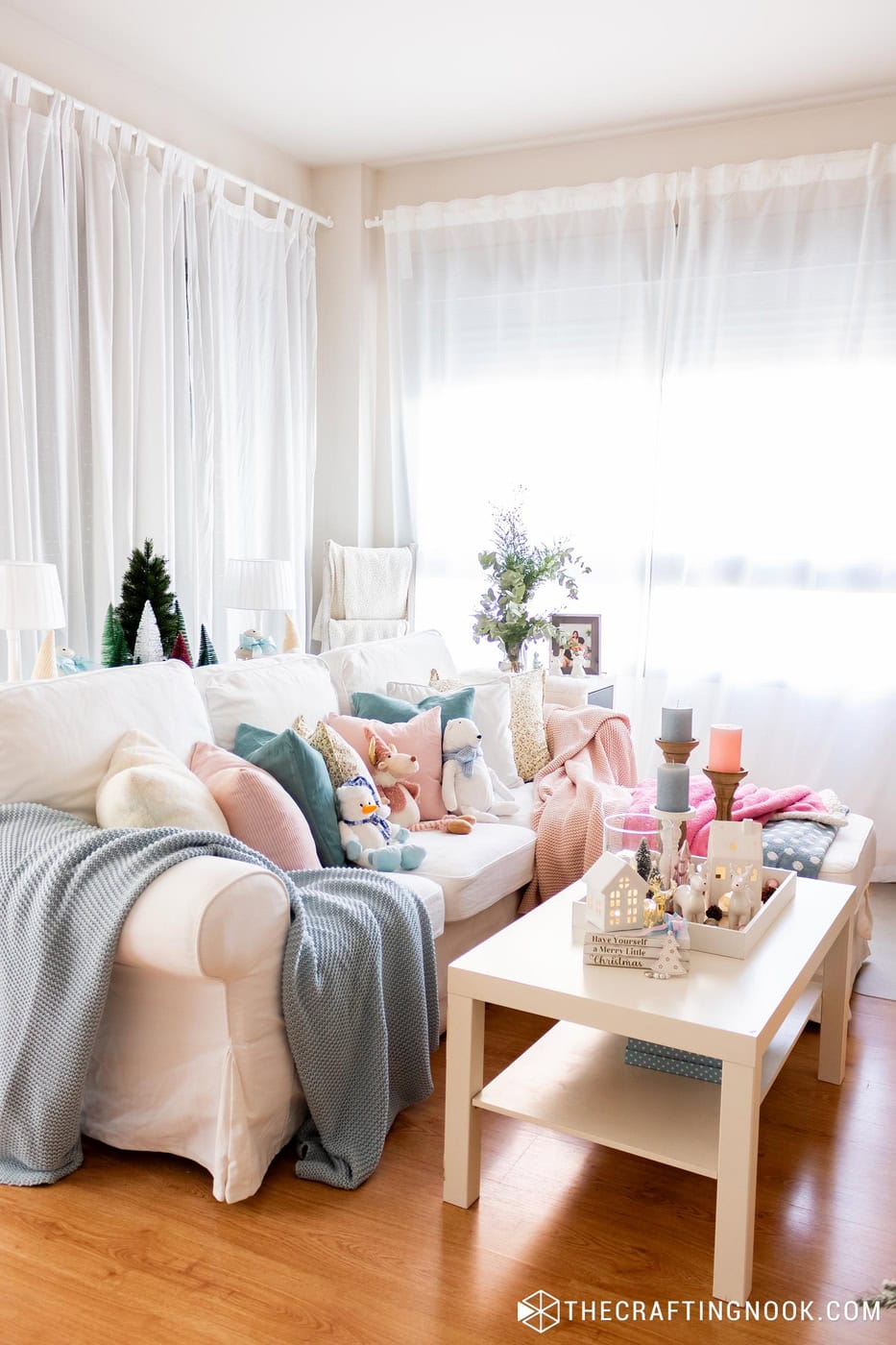 White sofa with blue and pink blankets, soft toys and a coffee table with a Christmas tray, in a bright room with sheer curtains