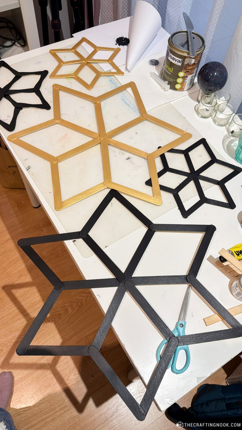 Large and small popsicle stick snowflakes painted black and gold arranged on a craft table.