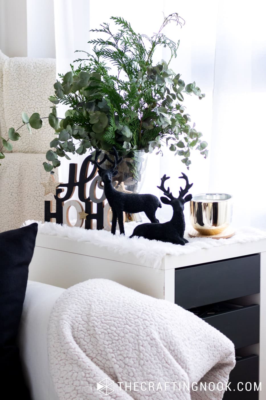 Christmas side cabinet decorated with eucalyptus greenery, black reindeer, festive lettering, and white faux-fur accents.
