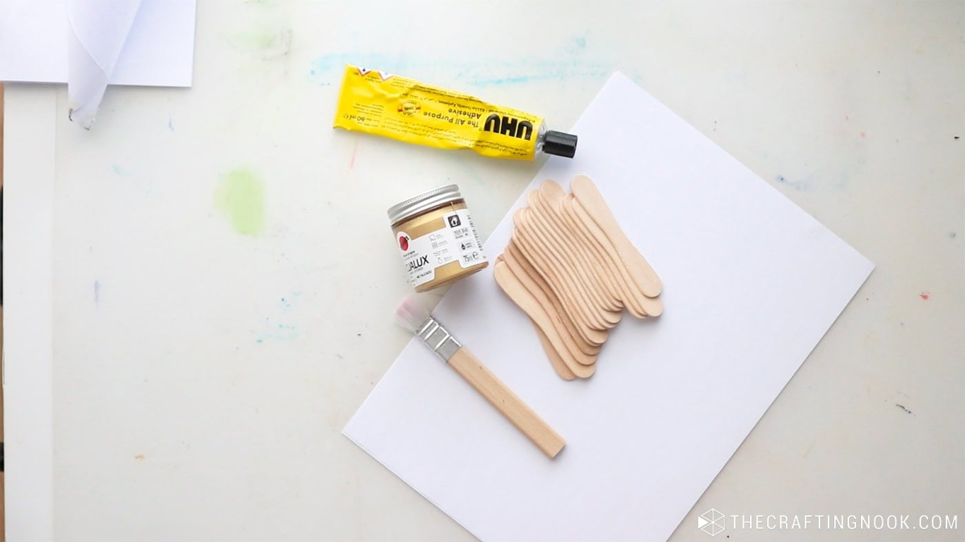 Supplies for making DIY popsicle stick snowflakes, including wooden craft sticks, UHU glue, metallic gold acrylic paint, a paintbrush, and protective paper.