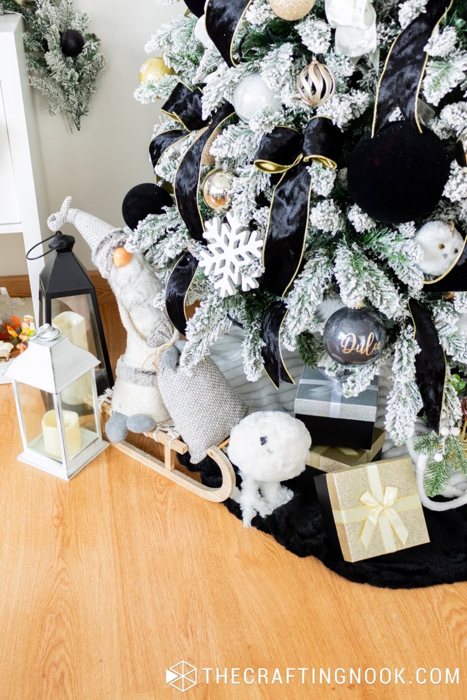 Christmas gnome sitting on a wooden sled beside a flocked tree decorated with black velvet ribbons, gold ornaments, lanterns and wrapped gifts.
