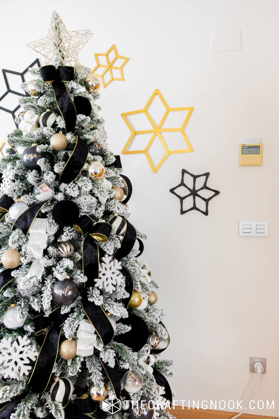 Upper section of a flocked Christmas tree with a gold wire star topper and handmade black and gold popsicle stick snowflakes on the wall.