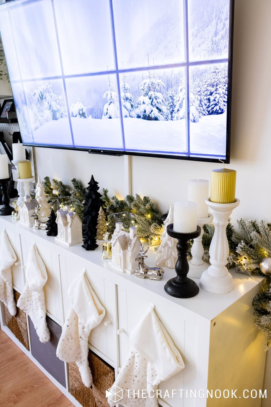 Christmas TV console with illuminated flocked garland, black and white ceramic houses, gold accents, and white stockings.
