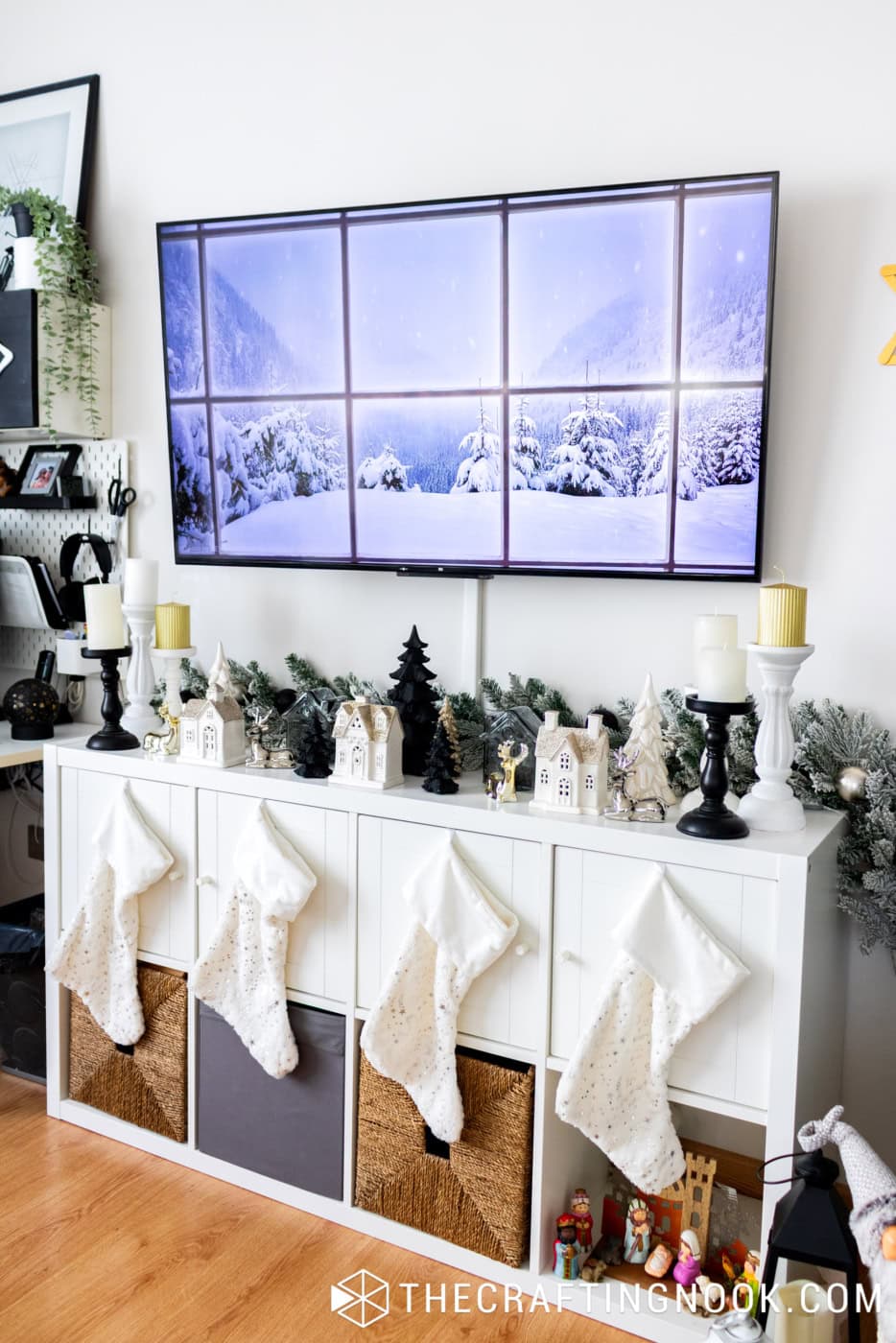 White TV console decorated with black and gold Christmas village, snowy greenery, four white stockings, and a winter scene on the television.

