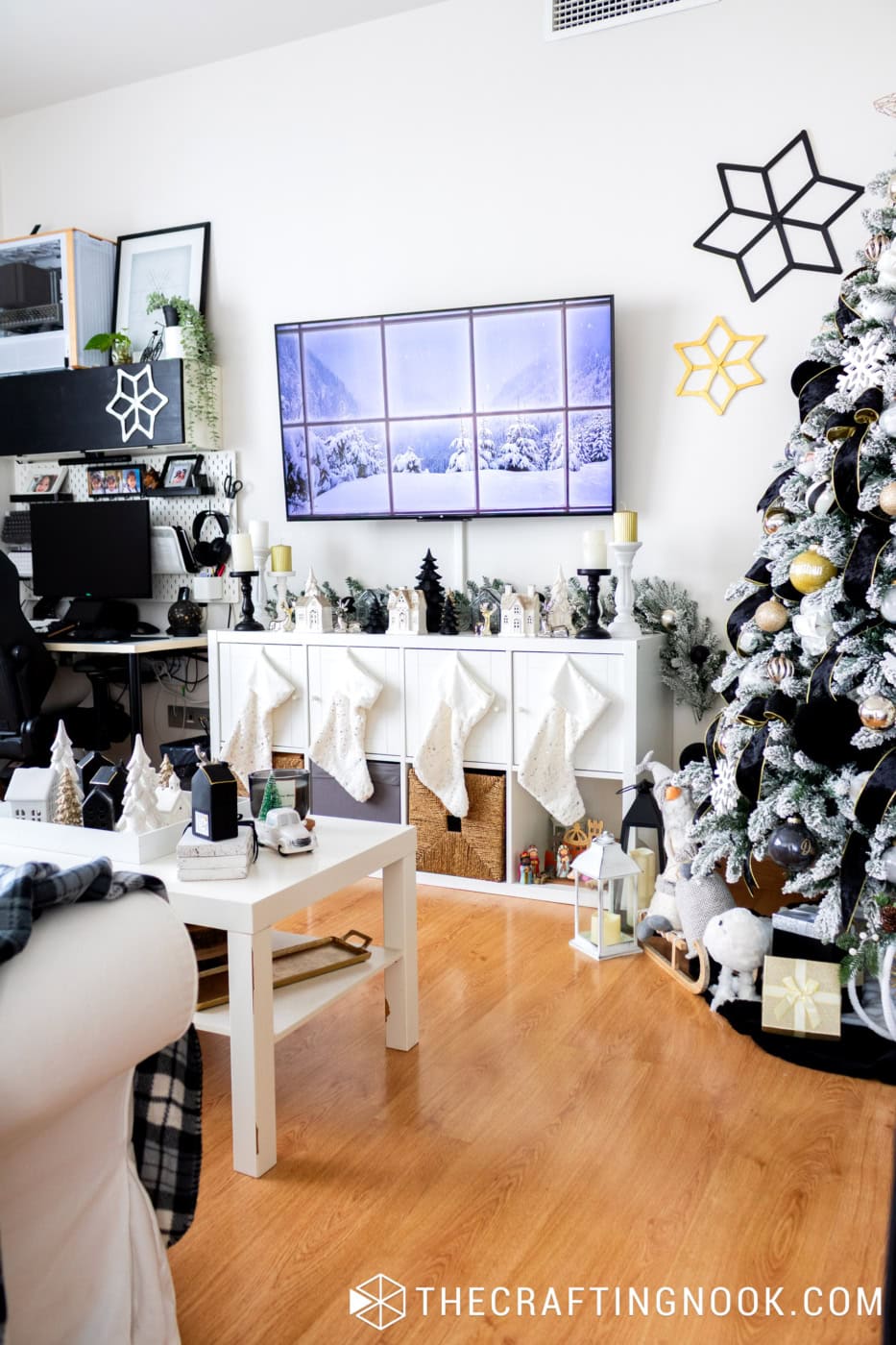 Black, white, and gold Christmas living room with a decorated TV console, white stockings, coffee table village, and flocked Christmas tree.
