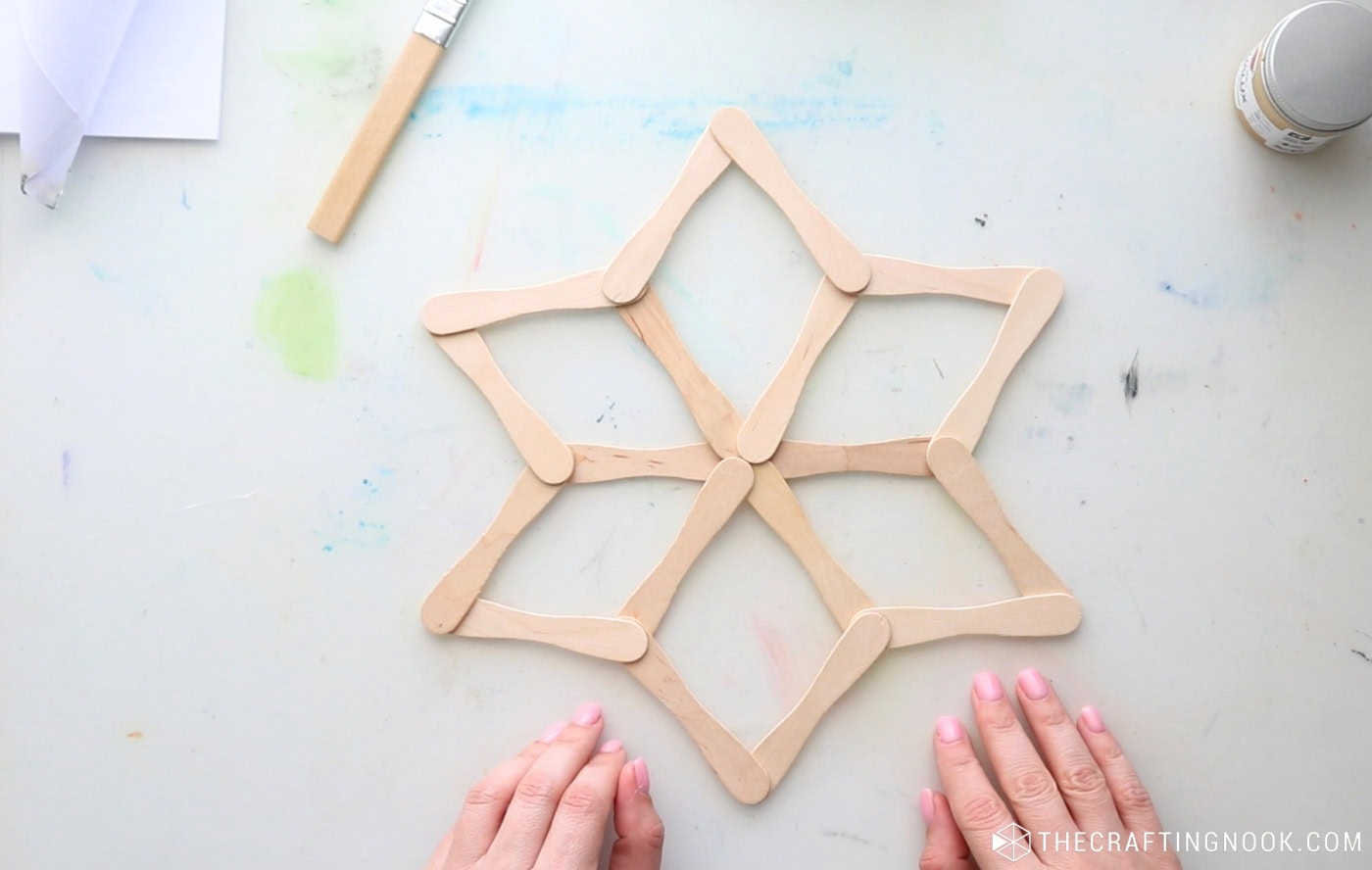 Unpainted popsicle stick snowflake with six geometric points assembled and ready for painting.