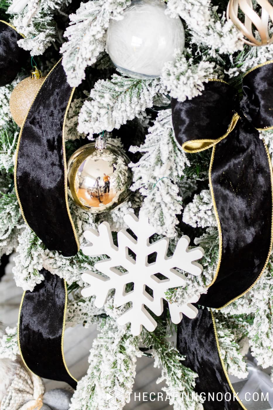 White wooden snowflake ornament hanging among snowy Christmas tree branches, black velvet ribbons with gold trim, and shiny gold baubles.